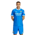 A man posing in a bright blue Real Madrid soccer kit with white Adidas and Emirates logos, standing against a solid black background.