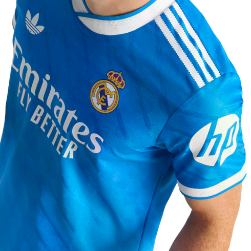 A blue Real Madrid soccer jersey with white Adidas Trefoil, Emirates, and HP logos, white shoulder stripes, and the team's crest.