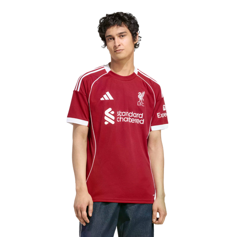 A young man modeling a red Liverpool FC jersey with white trim and sponsor logos, set against a solid black background.