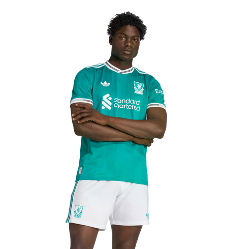 A man models a teal Liverpool FC soccer jersey with white trim and matching white shorts, featuring the Adidas Originals and Standard Chartered logos against a black background.