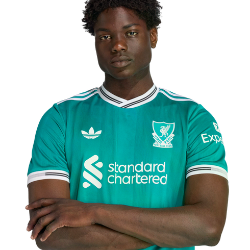 A young Black man with arms crossed wearing a teal Liverpool FC soccer jersey against a black background.