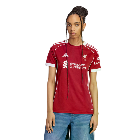 A woman with long braids models a red Liverpool F.C. soccer jersey with white trim and sponsor logos, paired with blue jeans against a black background.