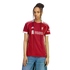 A woman with long braids models a red Liverpool F.C. soccer jersey with white trim and sponsor logos, paired with blue jeans against a black background.