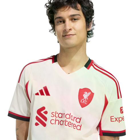 A young man with dark wavy hair wearing a white and red Liverpool FC soccer jersey, smiling against a plain black background.