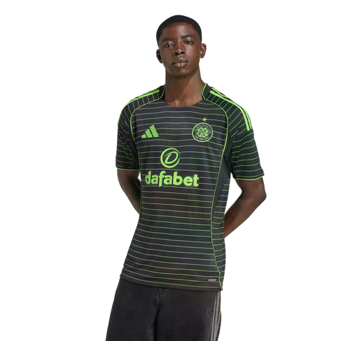 A young man models a black Celtic FC jersey featuring thin neon green horizontal stripes and logos, posed against a solid black background.