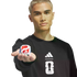 A blonde man in a black Adidas FIFA 2026 sweatshirt holds out a small white cube with red and blue branding against a black background.