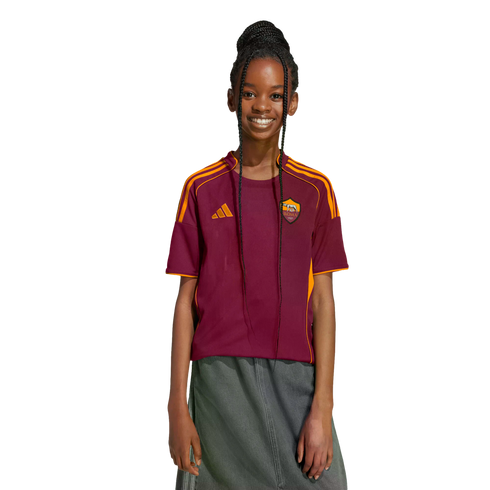 A young person with long braids smiles, wearing a maroon and orange AS Roma jersey and a grey skirt against a black background.