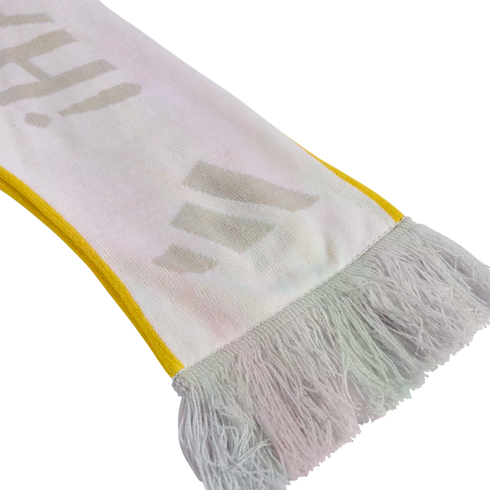 A close-up of a white knitted scarf with yellow edges and white tassels, featuring faint grey letters printed on the fabric.