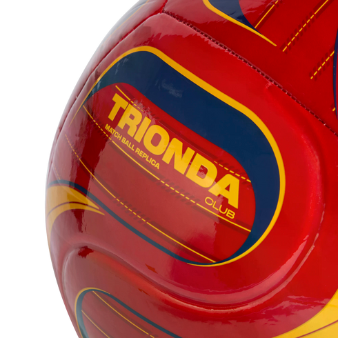 Close-up of a red soccer ball with yellow and blue graphics, featuring the yellow text "TRIONDA CLUB MATCH BALL REPLICA" against a black background.