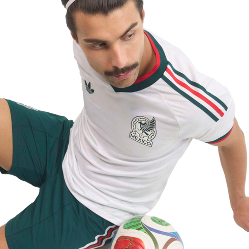 A man with a mustache wearing a white Mexico national soccer team jersey and green shorts sits with a soccer ball at his feet against a black background.