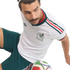 A man with a mustache wearing a white Mexico national soccer team jersey and green shorts sits with a soccer ball at his feet against a black background.
