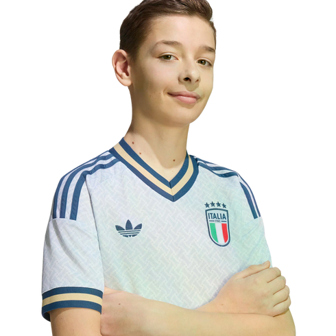 A young boy smiles with arms crossed, wearing a white Adidas Italian national soccer team jersey against a solid black background.