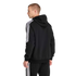 Rear view of a man wearing a black Adidas tracksuit with three white stripes on the sleeves and legs, looking to the side against a plain light grey background.