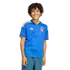 A smiling boy with curly hair wearing a blue patterned Italy soccer jersey and blue jeans, standing against a black background.