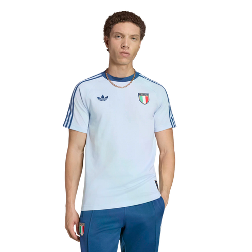 A young man with curly brown hair wearing a light blue Adidas Italy national team t-shirt and dark blue track pants, posed against a solid black background.