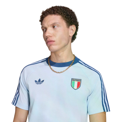 A portrait of a young man with curly brown hair looking away against a black background. He wears a light blue Adidas t-shirt featuring dark blue shoulder stripes, the Adidas logo, the Italian national football team crest, and a chunky gold chain.