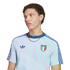 A portrait of a young man with curly brown hair looking away against a black background. He wears a light blue Adidas t-shirt featuring dark blue shoulder stripes, the Adidas logo, the Italian national football team crest, and a chunky gold chain.