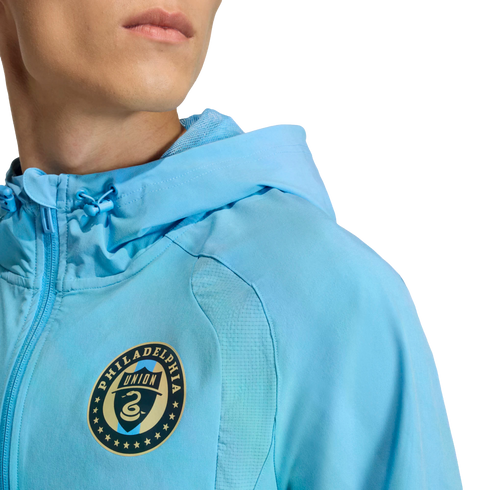 A person wears a light blue Philadelphia Union hooded jacket featuring the team's circular crest on the chest.