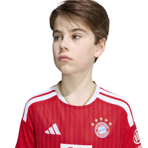 A young boy looking to the side while wearing a red and white FC Bayern Munich soccer jersey against a plain white background.