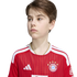 A young boy looking to the side while wearing a red and white FC Bayern Munich soccer jersey against a plain white background.