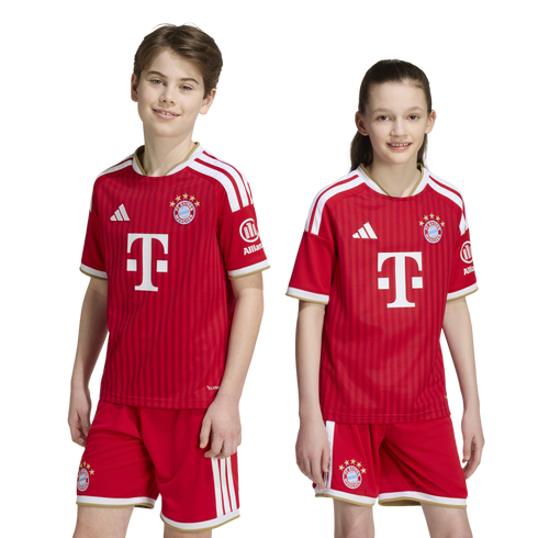 A boy and a girl smile wearing matching red FC Bayern Munich soccer uniforms, featuring the team's crest and sponsor logos, against a white background.
