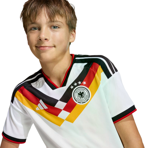 A smiling young boy with light brown hair wearing a white German national football team jersey featuring a black, red, and yellow graphic design.