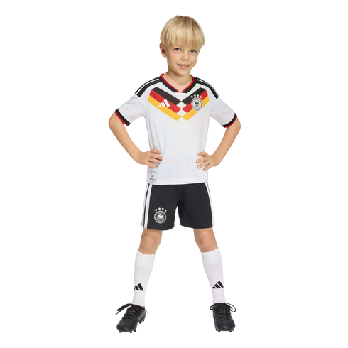 A smiling young boy wearing a Germany national team soccer kit, standing with hands on hips against a black background.