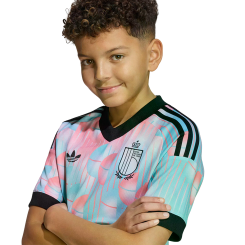 A smiling boy with curly hair poses with arms crossed, wearing a colorful pastel Belgian national football team jersey against a black background.
