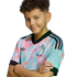 A smiling boy with curly hair poses with arms crossed, wearing a colorful pastel Belgian national football team jersey against a black background.