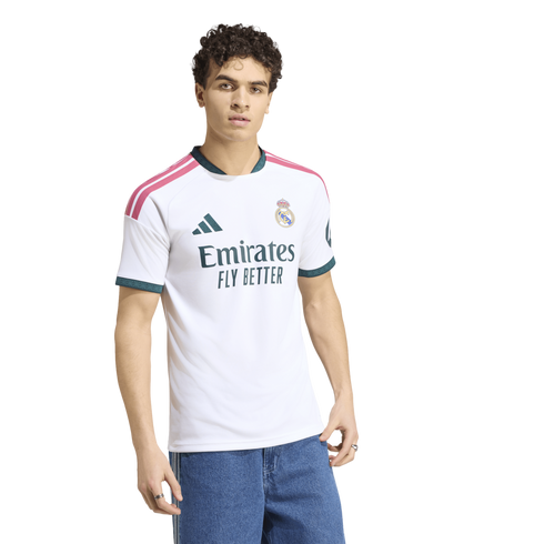 A young man with curly hair modeling a white Real Madrid home jersey and blue jeans against a white background.