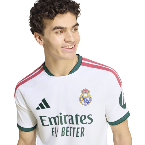 Smiling young man with curly hair wearing a white Real Madrid soccer jersey with green and pink accents.