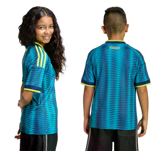 A girl and boy wearing teal and navy patterned Colombia soccer jerseys with yellow trim. One smiles at the camera; the other shows the back view with "COLOMBIA" on the neck.