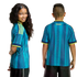 A girl and boy wearing teal and navy patterned Colombia soccer jerseys with yellow trim. One smiles at the camera; the other shows the back view with "COLOMBIA" on the neck.
