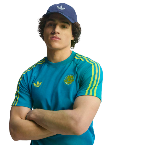 A man in a blue Adidas cap and teal soccer t-shirt with yellow stripes, posing with his arms crossed against a black background.