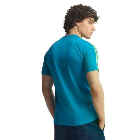 Back view of a man with curly hair in a teal t-shirt with yellow sleeve stripes, looking over his shoulder against a black background.