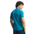 Back view of a man with curly hair in a teal t-shirt with yellow sleeve stripes, looking over his shoulder against a black background.