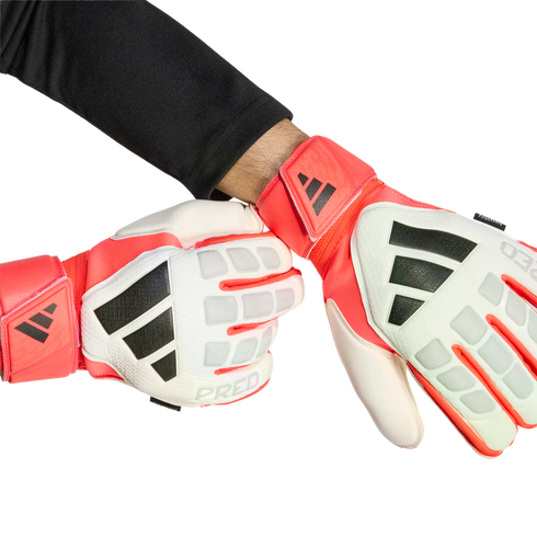 A person wears red, white, and black Adidas Predator goalkeeper gloves against a solid black background. One hand is clenched in a fist, and the other is open.