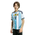 A young person with brown hair smiling in an Argentina national football team jersey with the number 10 and three stars, against a solid black background.