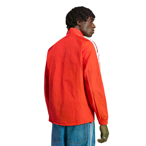Rear view of a man with dreadlocks wearing a bright orange zip-up jacket and blue jeans against a black background.