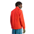 Rear view of a man with dreadlocks wearing a bright orange zip-up jacket and blue jeans against a black background.