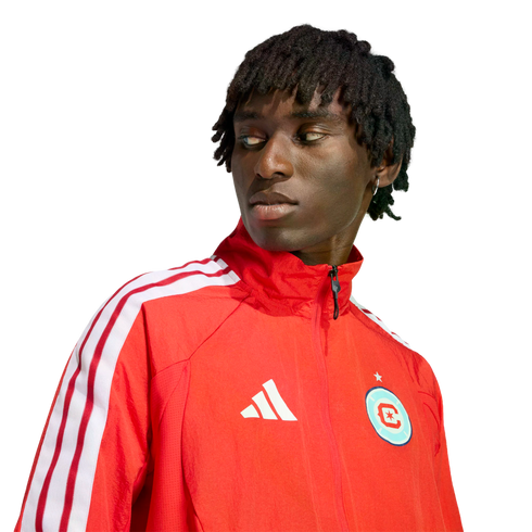 A man with short dreadlocks wearing a red Adidas track jacket with white stripes, looking off-camera against a black background.