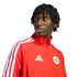 A man with short dreadlocks wearing a red Adidas track jacket with white stripes, looking off-camera against a black background.