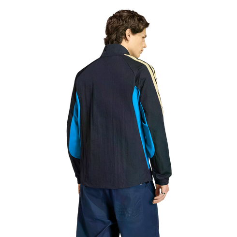 Rear view of a person in a black track jacket with blue side panels and cream stripes, wearing navy blue pants, against a black background.