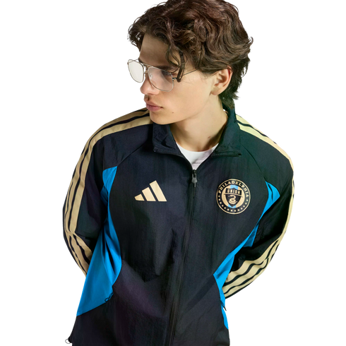 Young person with wavy hair and glasses wearing a black, blue, and gold Philadelphia Union Adidas track jacket against a black background.
