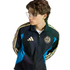 Young person with wavy hair and glasses wearing a black, blue, and gold Philadelphia Union Adidas track jacket against a black background.