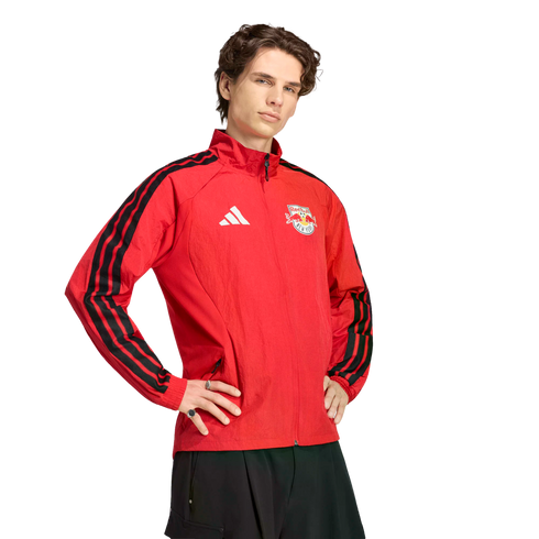 A young man wearing a red New York Red Bulls Adidas track jacket with black stripes, posing with hands on hips against a black background.