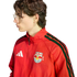 Side profile of a young person with glasses wearing a red Adidas New York Red Bulls track jacket against a black background.