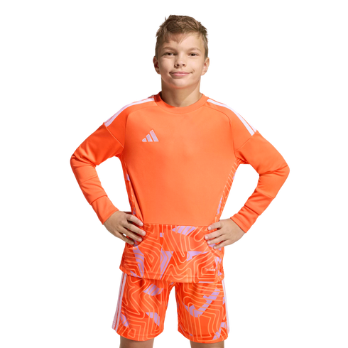 Boy in an orange long-sleeved Adidas soccer kit with a geometric pattern, posing with hands on hips against a solid black background.