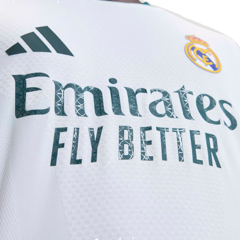 Close-up of a white Real Madrid soccer jersey featuring the Adidas logo, club crest, and "Emirates FLY BETTER" sponsor text with a graphic pattern inside the letters.