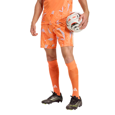 A person from the waist down wearing an orange patterned soccer kit and black cleats, holding a soccer ball against a solid black background.